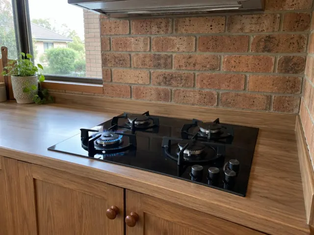 Black gas cooktop on a timber-look benchtop with brick splashback in a home kitchen in Chisholm