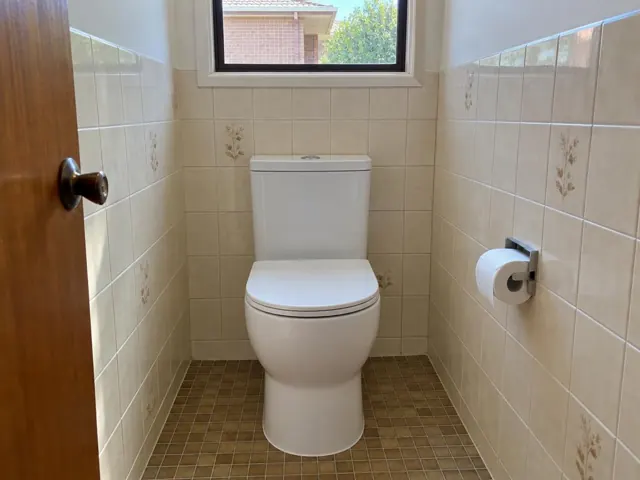 White toilet in a tiled small bathroom with a window and roll holder in Gillieston Heights